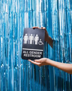 Eine Person hält ein Toiletten-Schild für Unisex-Toiletten vor blauem Hintergrund in der Hand