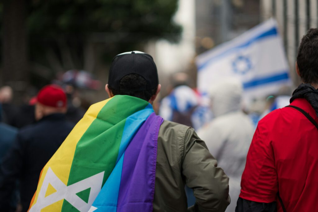 Eine Person steht mit dem Rücken zur Kamera und trägt eine Regenbogenflagge mit Davidstern.