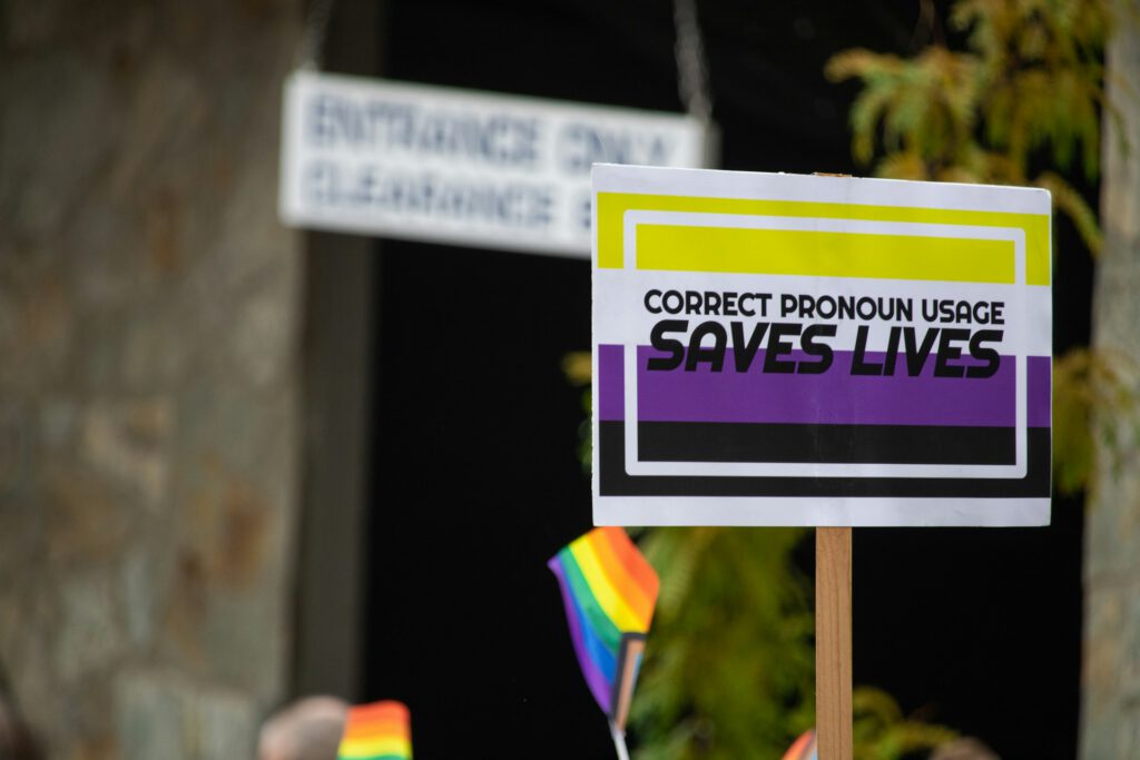 Auf einer CSD-Parade wird ein Schild in den Farben gelb, weiß, violett, schwarz und mit der Aufschrift "Correct Pronoun Usage Saves Lives" hochgehalten.