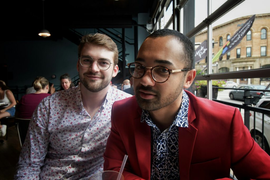 Zwei junge Männer sitzen nebeneinander in einem Café. (Symbolfoto für Artikel "Nach Aufregung über Wahlumfrage zur Bundestagswahl 2025 – jetzt spricht Planetromeo")