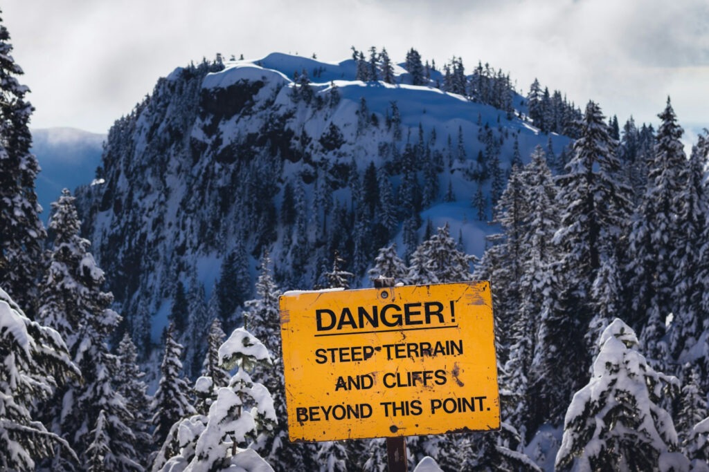 Blick auf verschneites Gebirge, im Vordergrund ein Warnschild, auf dem vor steilen Abhängen gewarnt wird.