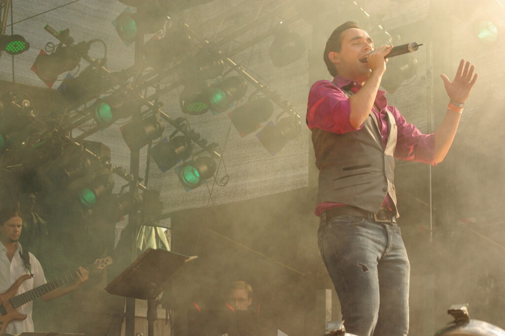 Daniel Küblböck singt auf der Bühne des CSD Köln 2009. Küblböck trägt ein pinkes Hemd, eine graue Weste und Hose und hält ein Mikrofon in der Hand.