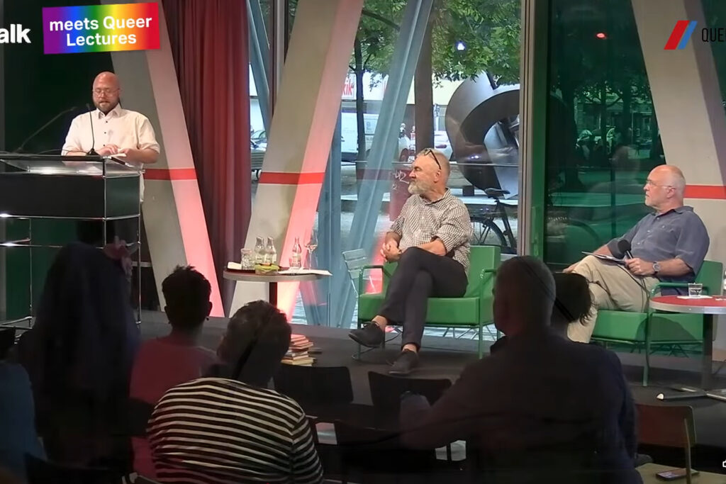 Till Randolf Amelung steht am Pult auf der Bühne der taz Kantine und hält seine Laudatio auf das Porträt von Hertha Wind. Daneben sind von links nach rechts sitzend Rainer Nicolaysen und Jan Feddersen. 