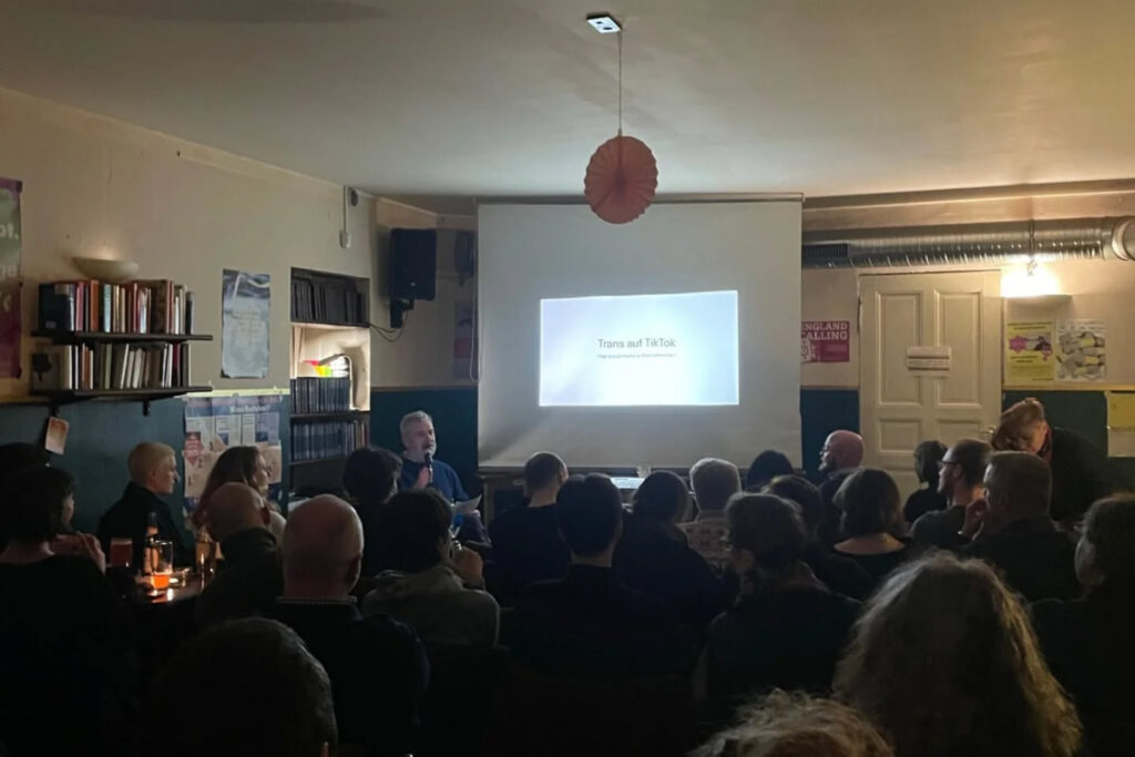 Jan Feddersen sitzt im Bajszel vor einer Leinwand, in der auf einer Powe-Point-Folie das Vortragsthema genannt wird (Foto: Privat).