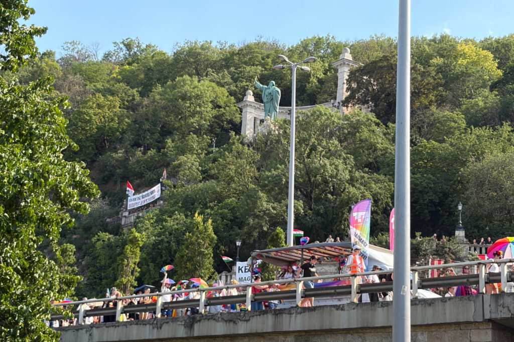 Budapest Pride: Eine große Menschenmenge zieht über eine Brücke, viele halten bunte Regenschirme und Regenbogenflaggen wie bei einer Pride‑Parade. Hinter der Brücke erhebt sich ein bewaldeter Hang, auf dem eine große Statue einer stehenden Figur mit ausgebreiteten Armen vor steinernen Säulen steht. Darüber hängen Banner und weitere Flaggen, der Himmel ist klar und blau.