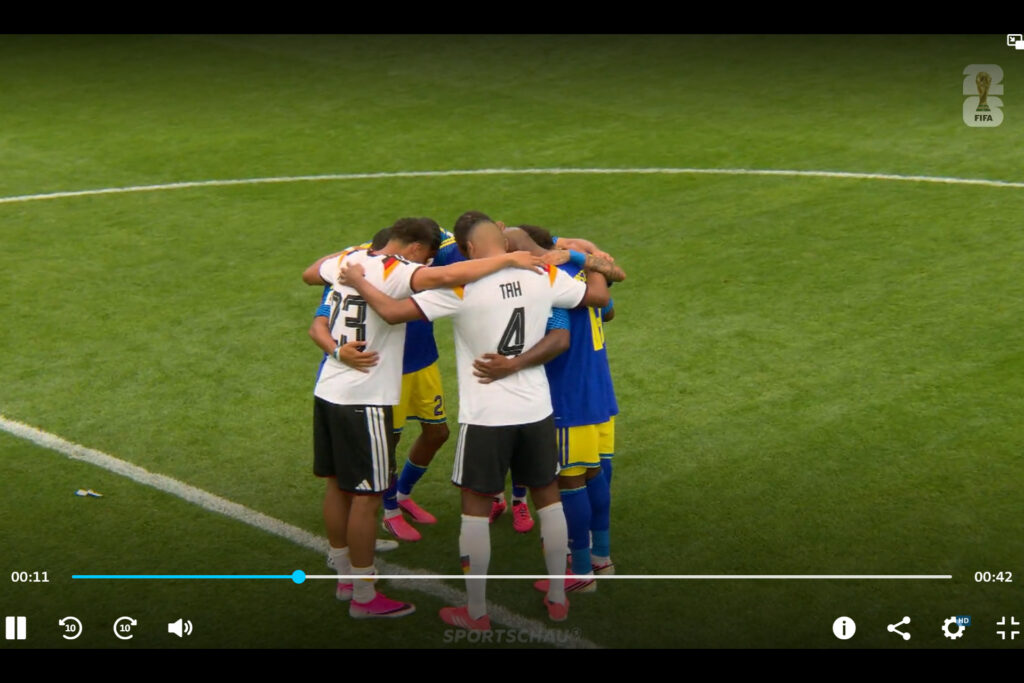 Die deutschen Nationalspieler Felix Nmecha und Jonathan Tah stehen mit Spielern von Curaçao auf einem Rasenplatz in einem engen Kreis mit den Armen umeinander. Zwei Spieler in weißen Trikots mit den Rückennummern 4 und 23 sind sichtbar, die anderen tragen blaue Trikots mit gelben Hosen. Die Szene ist ein Gebetskreis nach dem Spiel bei der WM 2026.