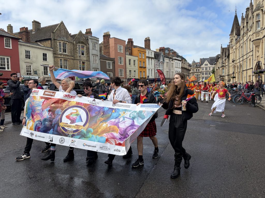 Eine bunte Gruppe von Menschen marschiert bei der Oxford Pride Parade, dem hiesigen CSD, durch eine historische Straße. Vorne tragen mehrere Personen ein großes, farbenfrohes Banner mit der Aufschrift „Oxford Pride Est. 2002 – 2026“, verziert mit regenbogenfarbenen Mustern und Logos verschiedener Sponsoren. Hinter ihnen laufen weitere Teilnehmende in leuchtenden Outfits, einige mit Pride‑Flaggen. Die Stimmung ist fröhlich und festlich, eingerahmt von alten Gebäuden im Hintergrund.