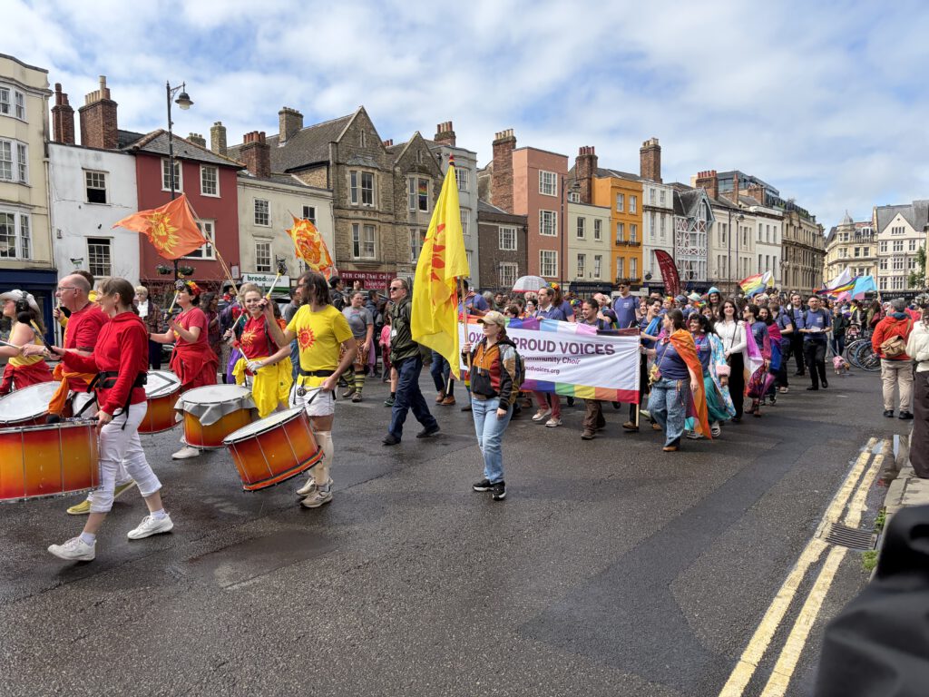 Eine Pride‑Parade zieht durch eine historische Straße, der Boden ist vom Regen noch nass. Vorne spielen Menschen in roten und gelben Outfits große Trommeln und tragen orange‑gelbe Fahnen mit Sonnensymbolen. Dahinter marschiert eine größere Gruppe mit einem Banner, auf dem „Oxford Proud Voices – Oxford’s LGBTQ+ community choir“ steht. Viele Teilnehmende tragen bunte Kleidung und Regenbogenflaggen, die Stimmung wirkt fröhlich und lebendig.