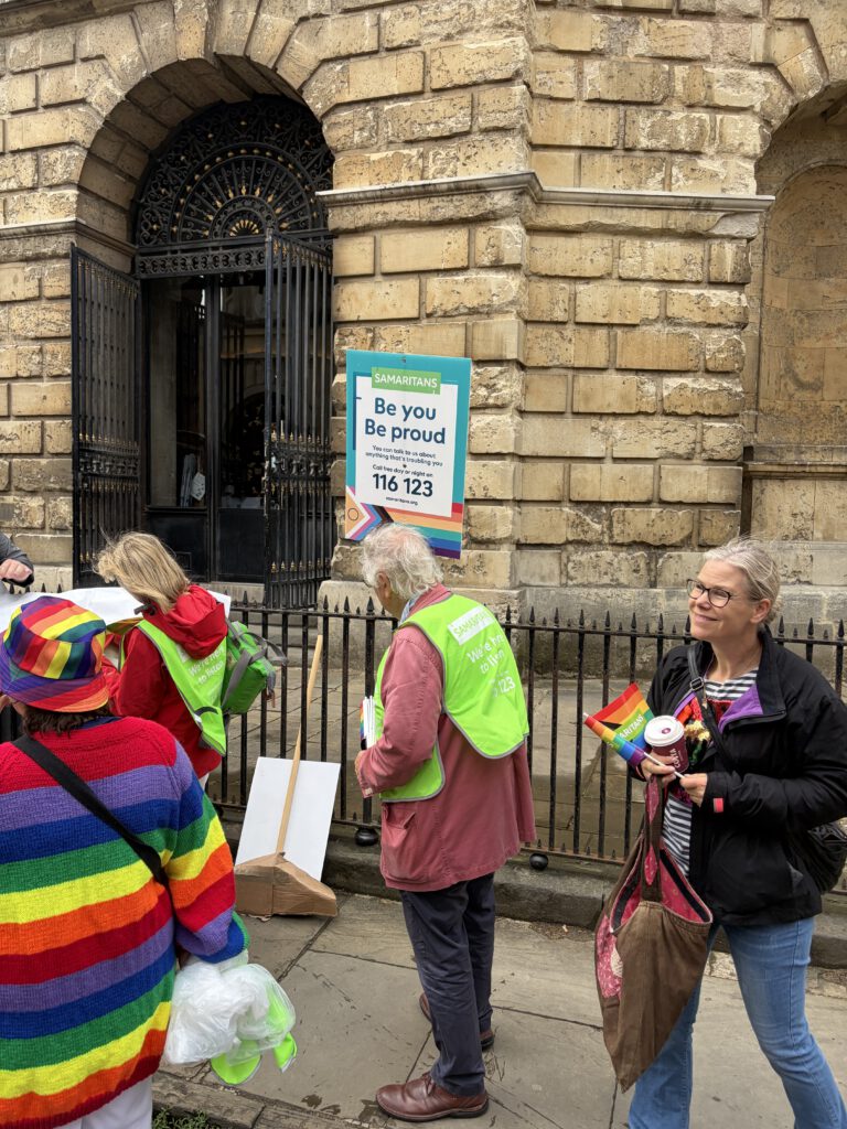 Mehrere Personen stehen auf einem gepflasterten Gehweg vor einem alten Steingebäude mit verziertem schwarzen Eisentor. Einige tragen leuchtend grüne Westen mit der Aufschrift „Samaritans“, eine Person trägt eine regenbogenfarbene Jacke und Mütze. Mehrere Regenbogenflaggen sind sichtbar. An der Wand hängt ein farbenfrohes Schild mit der Botschaft „Be you. Be proud.“ sowie einem Hinweis auf die kostenlose Samaritans‑Telefonnummer. Die Szene wirkt wie ein unterstützendes, inklusives Angebot im Rahmen eines Pride‑Events.