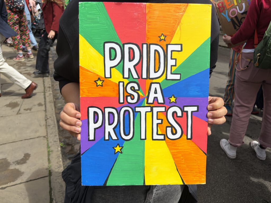Eine Person hält ein Schild mit regenbogenfarbenem Hintergrund hoch, darauf steht in großen schwarzen Buchstaben „Pride is a protest“, verziert mit kleinen gelben Sternen. Im Hintergrund sind weitere Menschen auf einer Straße zu sehen, die an einer Demonstration oder Pride‑Veranstaltung teilnehmen.