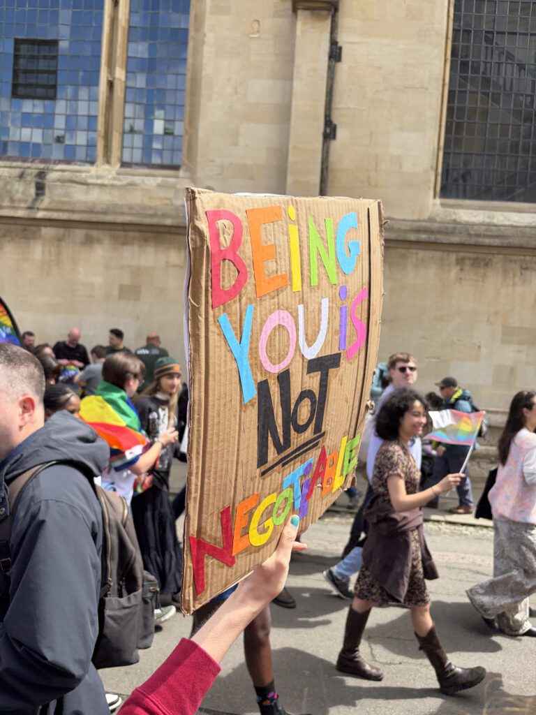 Eine Person hält ein Pappschild mit der bunten Aufschrift „Being you is not negotiable“ in die Höhe. Hinter ihr laufen mehrere Menschen mit Regenbogenflaggen eine Straße entlang, vorbei an einem großen Steingebäude mit hohen Fenstern. Die Szene wirkt wie ein Pride‑Marsch, der Selbstbestimmung und queere Sichtbarkeit feiert.