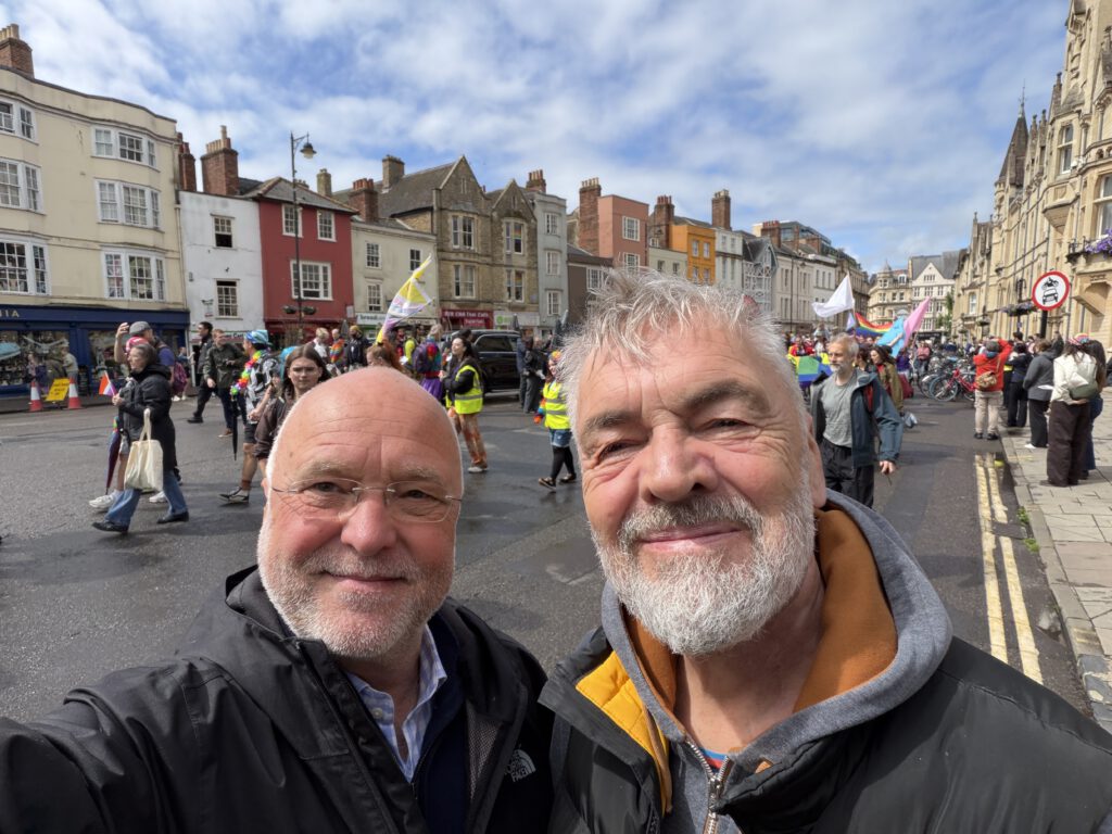Rainer Nicolaysen (links) und Jan Feddersen (rechts) stehen dicht beieinander auf einer Straße und machen ein Selfie. Hinter ihnen zieht eine bunte Parade vorbei, bei der Menschen mit farbenfrohen Fahnen und Bannern die Straße entlanglaufen. Die Umgebung besteht aus historischen Gebäuden mit verzierten Fassaden, und der Himmel ist teils bewölkt. Die Stimmung wirkt lebendig und festlich.