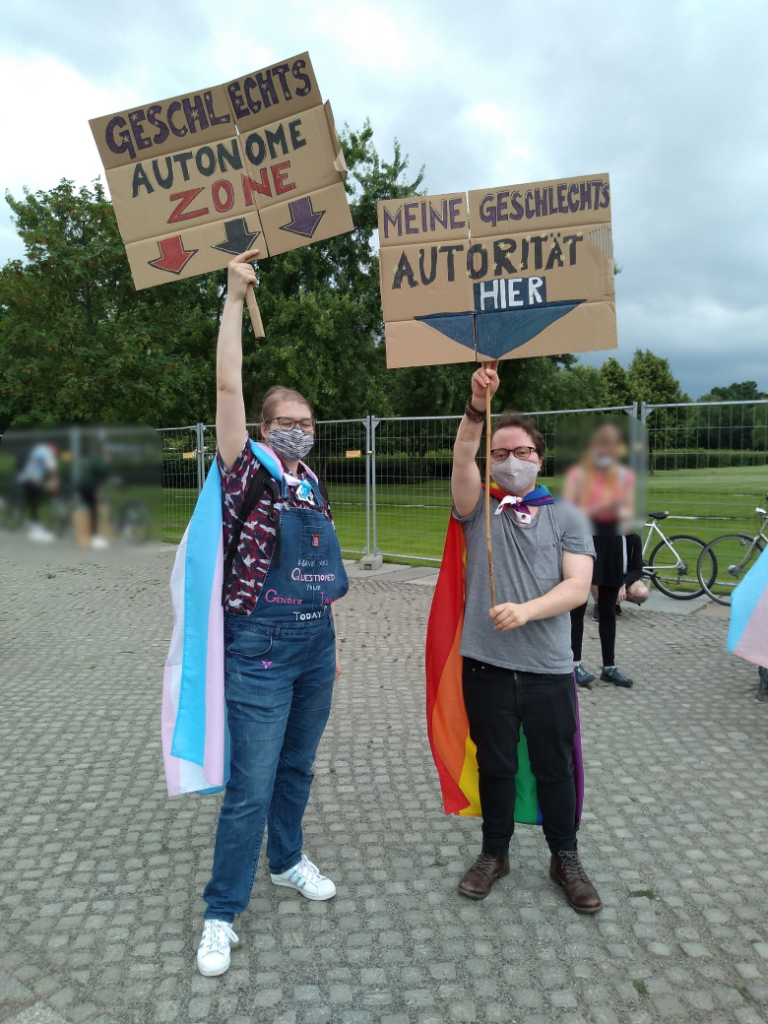 Zwei Teilnehmende an der Demo für das Selbstbestimmungsgesetz 2020 in Berlin vor dem Bundestag stehen nebeneinander. Die Person links hat eine Transflagge umgebunden und hält ein Schild hoch, auf dem steht "Geschlechtsautonome Zone". Die rechts neben ihr stehende Person hat eine Regenbogenflagge um den Hals geschlungen und hält ein SChild hoch, auf dem steht "Meine Geschlechtsautorität: Hier" Unter dem "Hier" ist ein Pfeil, der auf die Person selbst zeigt.