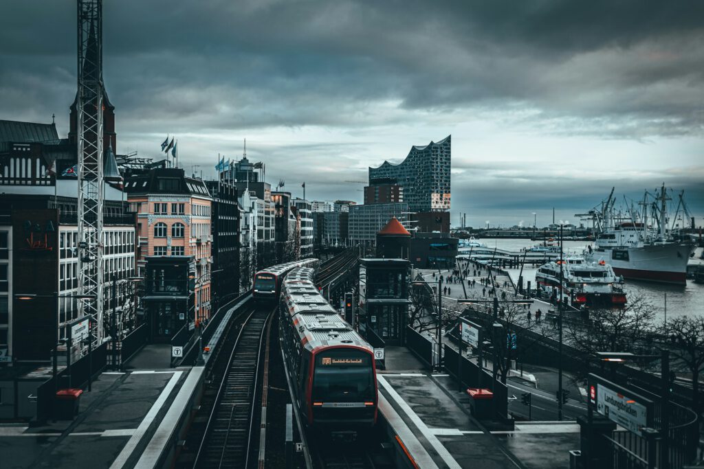 Ein erhöht verlaufender Bahnhof in Hamburg, einer europäischen Großstadt, auf dessen Gleisen zwei rote Züge nebeneinander stehen. Rundherum stehen mehrstöckige Gebäude, dahinter öffnet sich ein Hafen mit Kränen, Schiffen und Wasserflächen. Im Hintergrund ragt die markante Glasarchitektur der Hamburger Elbphilharmonie in den grauen, wolkigen Himmel. Symbolbild für Artikel
Zwei Entscheidungen der Justizministerkonferenz
