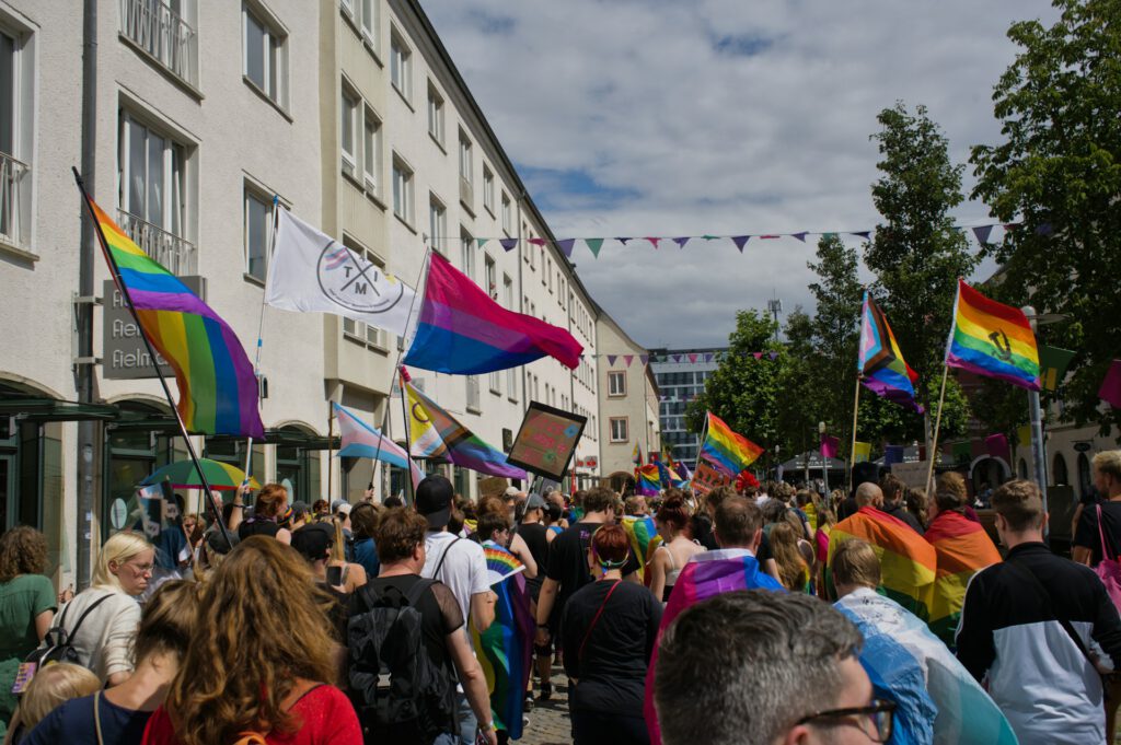 Sichtbarkeit: Eine große Menschenmenge zieht bei einer Pride‑Parade durch eine Straße einer kleineren Stadt. Viele Teilnehmende tragen Regenbogen‑, Bisexual‑ und Trans‑Pride‑Flaggen sowie Schilder mit Botschaften für LGBTIQ‑Rechte. Über der Straße hängen bunte Wimpelketten, und helle mehrstöckige Gebäude säumen den Weg.