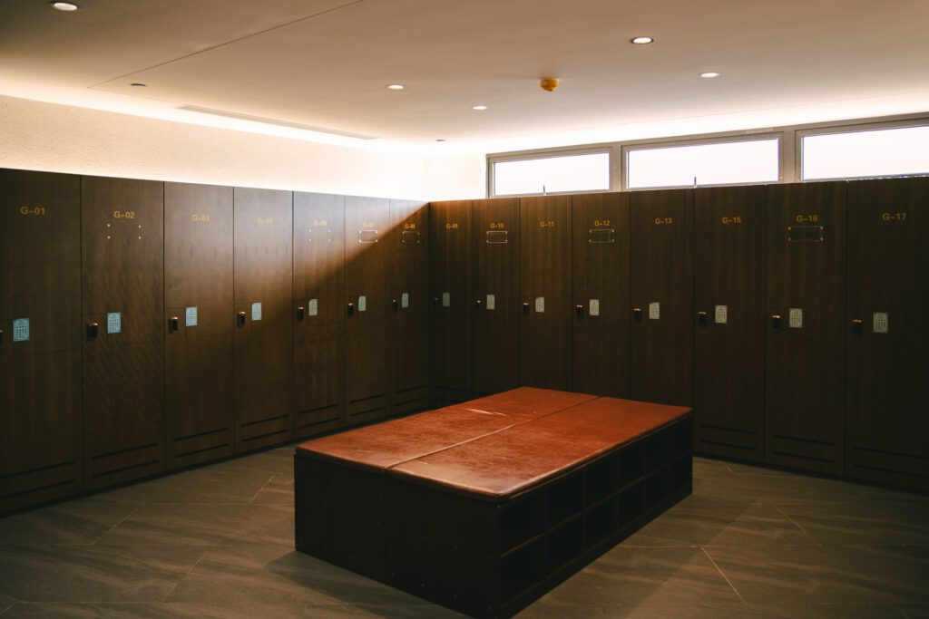 So stellt man sich die Gegebenheiten im Profifußball vor: Ein moderner Umkleideraum mit dunklen Holzschränken, die entlang zweier Wände im rechten Winkel angeordnet sind. Jeder Spind hat ein digitales Tastenfeld und gelbe Nummerncodes wie „G‑01“ bis „G‑17“. In der Raummitte steht eine große rechteckige Bank mit brauner Lederpolsterung und offenen Fächern darunter. Der Raum ist sauber, hell ausgeleuchtet und mit grauen Bodenfliesen gestaltet.