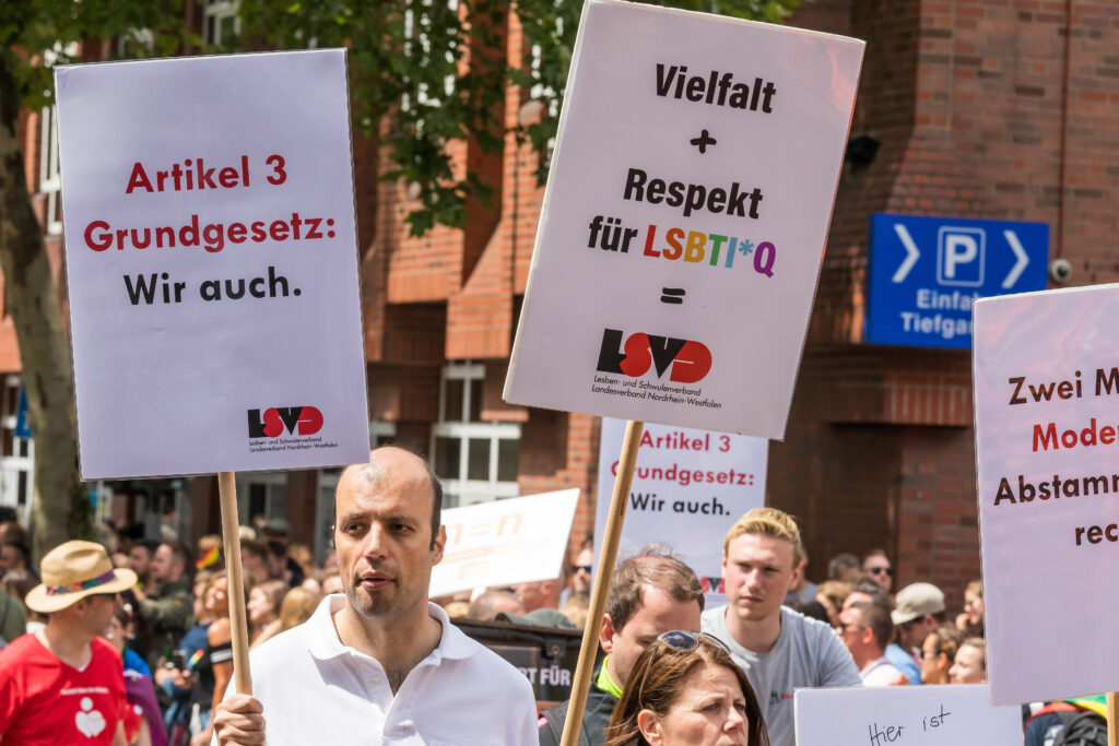 Eine Gruppe von Menschen zieht bei einer LGBTQ+-Demonstration über eine städtische Straße. Mehrere Teilnehmende halten Schilder hoch, darunter gut lesbar Artikel 3 Grundgesetz: Wir auch.“ sowie ein Plakat des LSVD mit dem Aufruf zu Vielfalt und Respekt für LSBTI*Q. Im Hintergrund stehen Backsteingebäude und ein Parkplatzschild, die Szene wurde beim CSD in Köln 2019 aufgenommen.