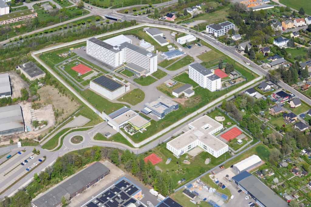 Hier darf ein Sexualstraftäter hinverlegt werden: Luftaufnahme des weitläufigen Gebäudekomplexes der JVA Chemnitz mit mehreren modernen, flach gedeckten Gebäuden, teils mit Solarpanels. Dazwischen liegen Grünflächen, Parkplätze und rot gefärbte Sportplätze. Der gesamte Bereich ist von Straßen umgeben und grenzt an Wohnhäuser sowie weitere Gewerbebauten.