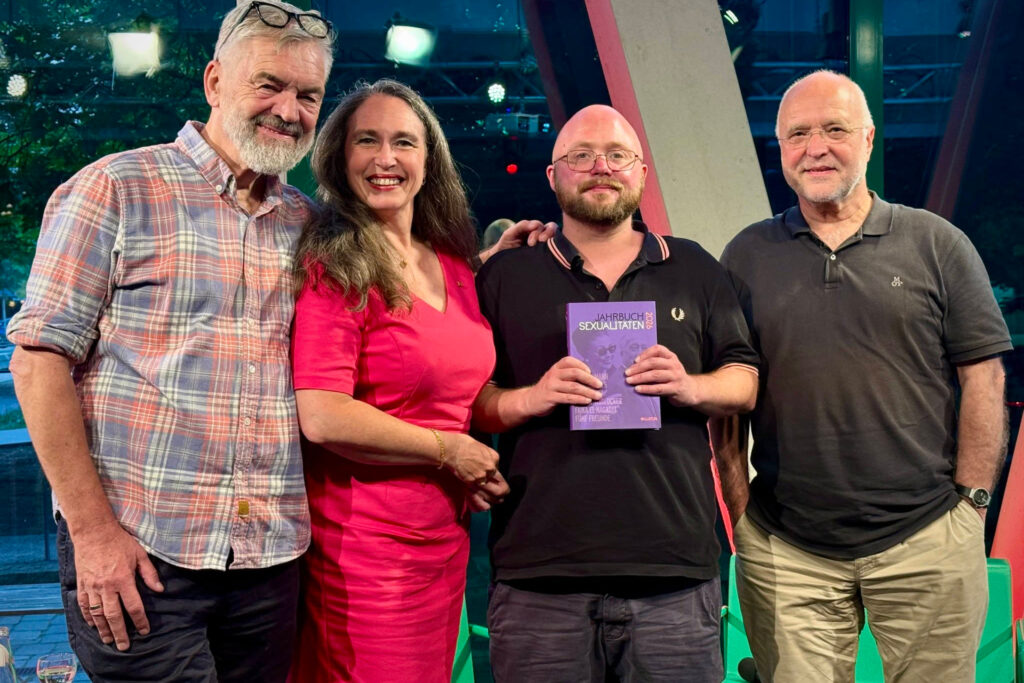 Alt‑Text: Jan Feddersen, Inge Bell, Till Randolf Amelung und Rainer Nicolaysen (von links nach rechts) stehen in einem modernen Innenraum nebeneinander und lächeln in die Kamera. Die Person zweitrechts hält ein lilafarbenes Buch mit dem Titel Jahrbuch Sexualitäten 2026 gut sichtbar vor sich. Hinter ihnen sind große Fenster mit Lichtreflexen und Metallstreben zu sehen, was auf eine Studio‑ oder Veranstaltungsumgebung hinweist.