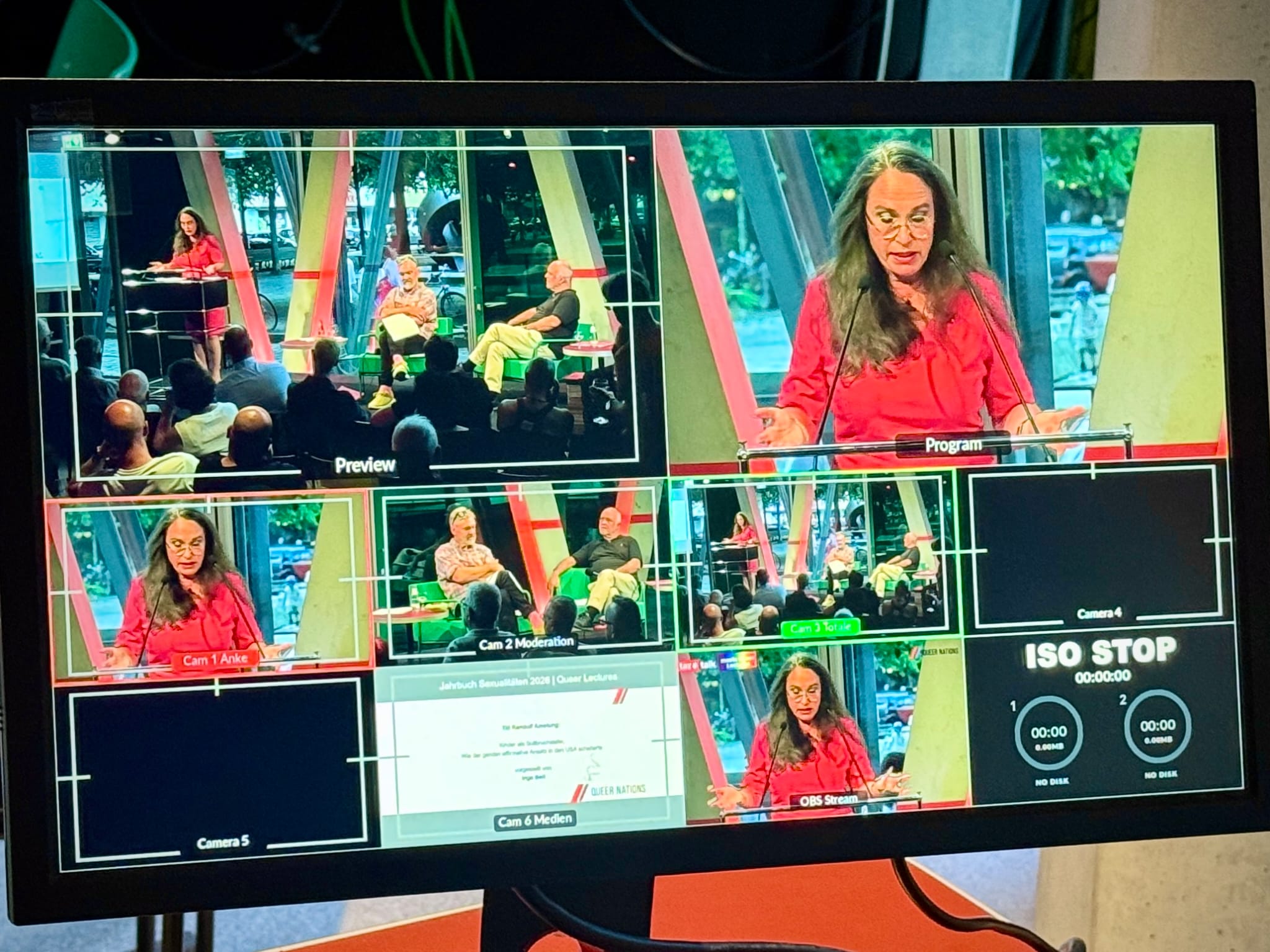 Ein Monitor zeigt die Benutzeroberfläche einer Video‑Produktion mit mehreren Kameraansichten eines Vortrags. Im Hauptbild („Program“) steht eine Person in roter Kleidung am Podium und spricht, während andere Fenster verschiedene Perspektiven auf Bühne und Publikum zeigen. Die Umgebung wirkt modern mit großen Fenstern und sichtbaren Stahlträgern.