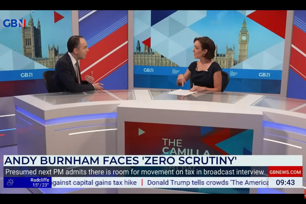 Ein Screenshot einer GB‑News‑Sendung zeigt Gesundheitsminister James Murray von der Labour-Party(links) und Host Camilla Tominey (rechts) an einem Nachrichtentisch im Studio, vor einer großen Hintergrundgrafik mit dem Londoner Parlament und Big Ben. Auf dem Bildschirm steht die Schlagzeile „Andy Burnham faces ‘zero scrutiny’“ sowie ein Untertitel über mögliche Steuerbewegungen. Am unteren Rand laufen Wetter‑ und Nachrichteneinblendungen, rechts ist die Uhrzeit 09:43 zu sehen.