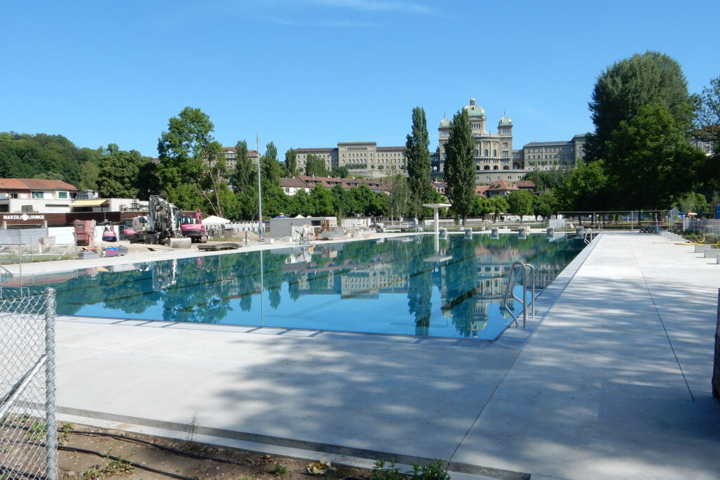 Ein Blick auf das frisch renovierte Außenbecken im Marzilibad in Bern mit klarem, türkisfarbenem Wasser, umgeben von einer breiten Betonfläche und Baugeräten. Im Hintergrund stehen historische Gebäude mit Kuppeldächern und reich verzierten Fassaden, eingerahmt von Bäumen unter einem wolkenlosen, blauen Himmel.