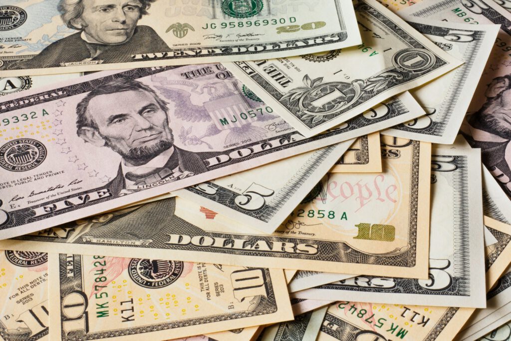 Ein verstreuter Stapel verschiedener US‑Dollar‑Banknoten – darunter 1‑, 5‑, 10‑ und 20‑Dollar‑Scheine – liegt übereinander. Die Porträts von Lincoln, Hamilton und Jackson sind sichtbar, die Farben und Details der Geldscheine heben sich klar voneinander ab. Symbolbild für Artikel Nicht nur in den USA: Profite mit Pubertätsblocker?