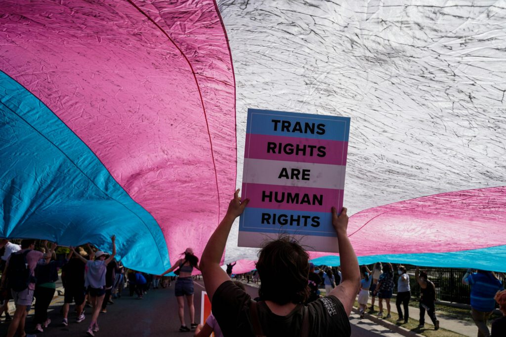 Eine Gruppe von Menschen nimmt an einer Demonstration teil und hält eine große Transgender‑Pride‑Flagge in Rosa, Blau und Weiß. Im Vordergrund steht eine Person mit einem Schild, auf dem „Trans Rights Are Human Rights“ steht. Symbolbild für Artikel "Zwischen Affirmation und Vorsicht: zur aktuellen medizinischen Debatte um Transkinder"