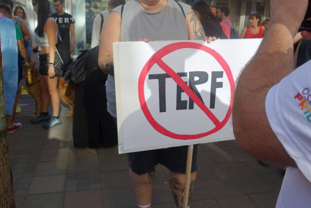 Eine Person hält bei der Pride Parade in Belgrad 2019 ein Schild hoch, auf dem das Wort Terf in schwarzen Buchstaben steht, durchgestrichen mit einem roten Kreis und Querbalken. Im Hintergrund sind weitere Menschen sowie ein Hund an der Leine zu sehen, die auf einer gepflasterten Fläche im Freien stehen.