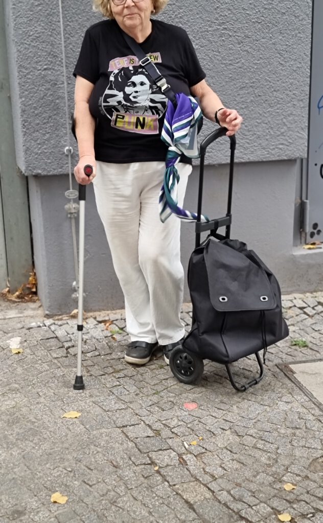 Eine ältere Person steht auf einem Kopfsteinpflaster neben einer grauen Wand. Sie hält in der rechten Hand einen Gehstock und in der linken einen schwarzen Einkaufsroller, an dessen Griff ein buntes Tuch befestigt ist. Die Person trägt ein schwarzes T‑Shirt mit grafischem Aufdruck und dem Text „TERF IS NEW PUNK“, dazu weiße Hose und dunkle Schuhe.
