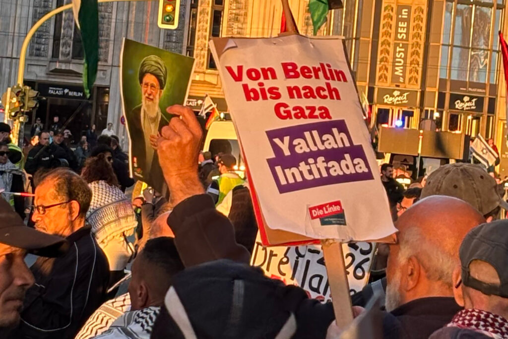 Demonstration "United for Palestine" am 10. Oktober 2026 in Berlin mit Teilnehmenden, Fahnen und einem Plakat mit der Aufschrift „Von Berlin bis nach Gaza – Yallah Intifada“. Auf dem Plakat ist das Logo der Partei "Die Linke" zu sehen - ein Ausdruck von israelbezogenem Antisemitismus. Daneben hält eine andere Person ein Bild mit dem im Februar 2026 bei einem israelischen Angriff getöteten Obersten Führer des Irans, Ali Chamenei, hoch. Im Hintergrund sind Polizei, weitere Protestierende und Gebäude der Innenstadt zu sehen.