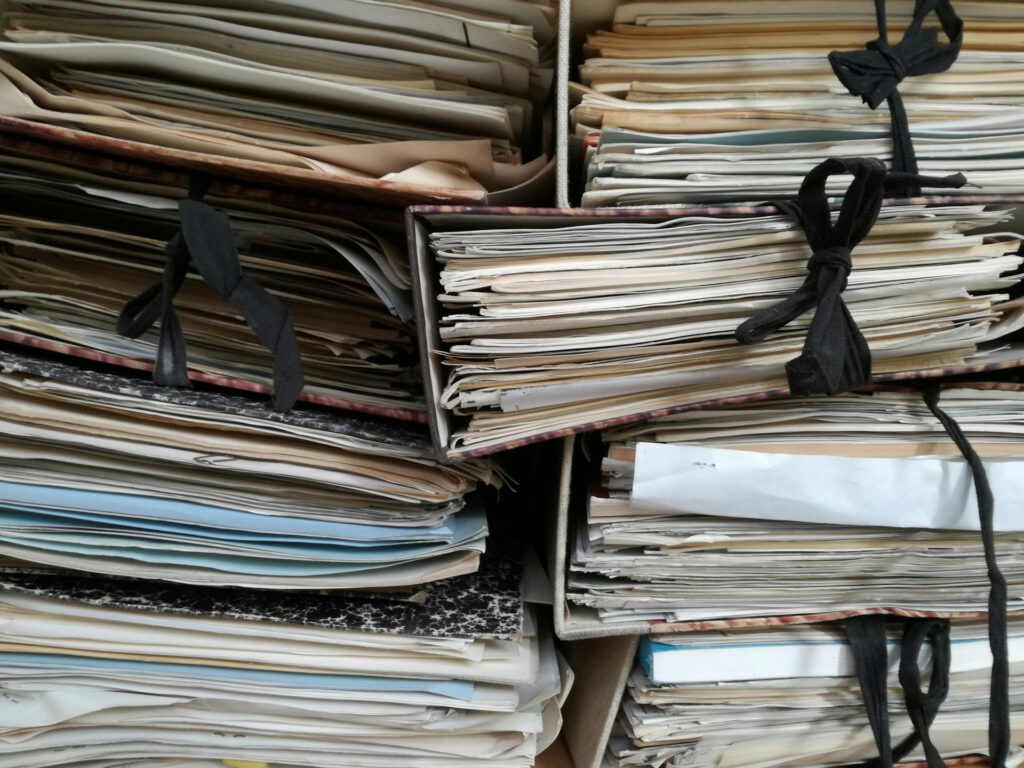 Mehrere dicht gestapelte Papierordner und Dokumentenmappen mit schwarzen Stoffbändern liegen übereinander. Die Mappen sind mit zahlreichen lose eingelegten, teils vergilbten Papierstapeln gefüllt. Das Bild vermittelt den Eindruck eines umfangreichen Papierarchivs. Symbolbild für Emanzipationshistorie mit blinden Flecken und fehlender Quellenbasis